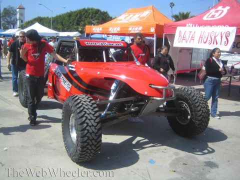 1990 Ford Bronco - red tube-frame off-road race buggy sits on pavement at an outdoor expo, with vendor tents and onlookers behind it.