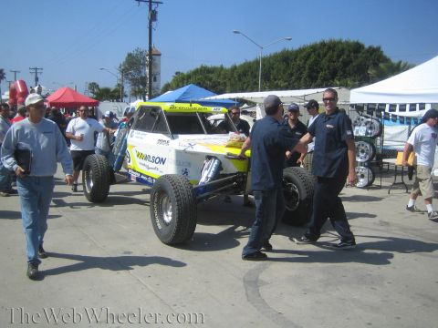 1990 Ford Bronco - Crew members maneuver a white and yellow sponsored off-road race buggy across pavement at a busy outdoor gathering.