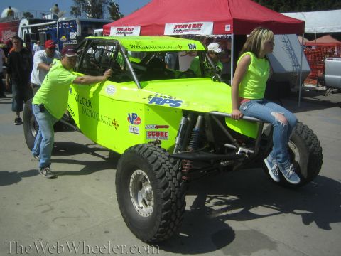 1990 Ford Bronco - neon green sponsored race buggy is pushed through an event while a young woman sits perched on its rear tire.