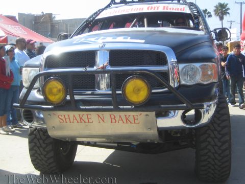 1990 Ford Bronco - front of a dark lifted Dodge Ram off-road truck shows yellow auxiliary lights and a bumper plate reading SHAKE N BAKE.