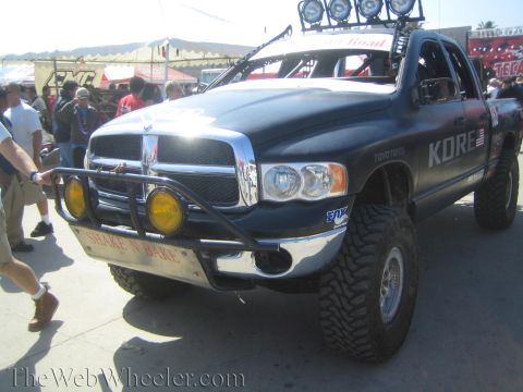 1990 Ford Bronco - lifted dark Dodge Ram off-road truck with a roof-mounted light bar and KORE graphics is shown at a crowded outdoor event.
