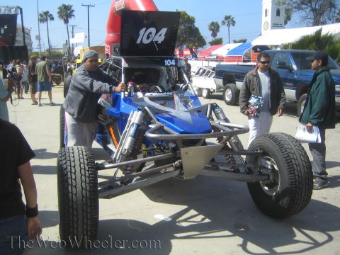 1990 Ford Bronco - man works on a blue single-seat off-road race buggy marked 104 on pavement at a sunny outdoor gathering.