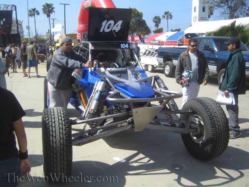 Blue single-seat off-road buggy number 104 with crew at outdoor event
