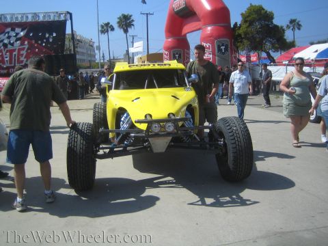 1990 Ford Bronco - yellow off-road race buggy sits near a tall red inflatable arch while spectators walk past at an outdoor event.