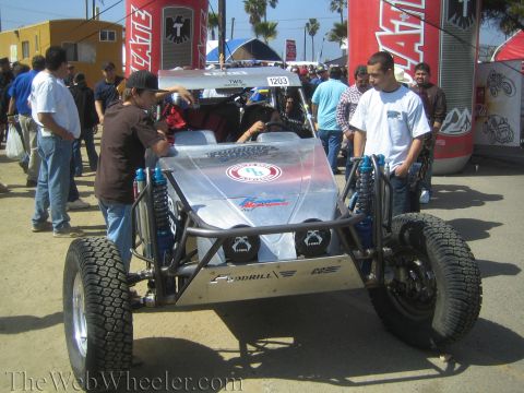 1990 Ford Bronco - bare aluminum-bodied off-road buggy marked 1203 is parked among a dense crowd beneath Tecate banners at an outdoor event.