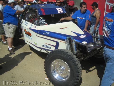 1990 Ford Bronco - Crew in matching blue shirts gather around a blue and white tube-frame off-road buggy on a dirt lot.