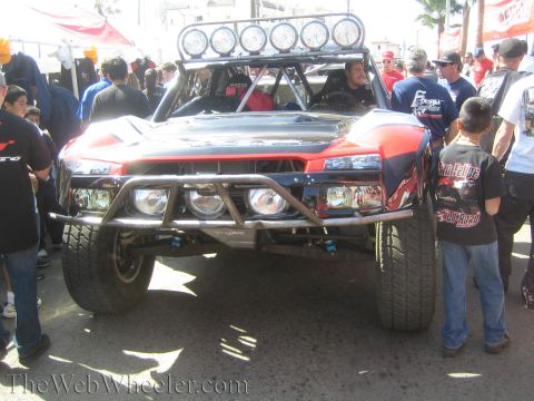 1990 Ford Bronco - front of a red and black off-road race truck with a six-light roof bar faces a crowd of spectators at an outdoor event.