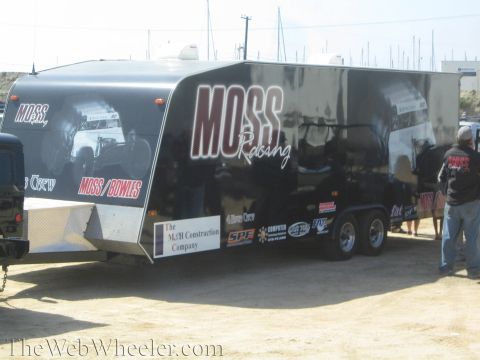 1990 Ford Bronco - black and silver dual-axle enclosed race trailer with Moss Racing graphics sits on a dirt lot as a man stands beside it.