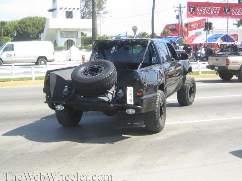 1990 Ford Bronco - black off-road prerunner truck with a bed-mounted spare tire drives down a street past Tecate banners at an event.