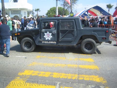 1990 Ford Bronco - black H1-style Hummer lettered POLICIA with a star badge is parked on a street crossed by yellow markings amid a crowd.