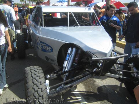 1990 Ford Bronco - silver-bodied tube-chassis off-road race car with an exposed front Fox coilover shock sits among spectators at an outdoor event.