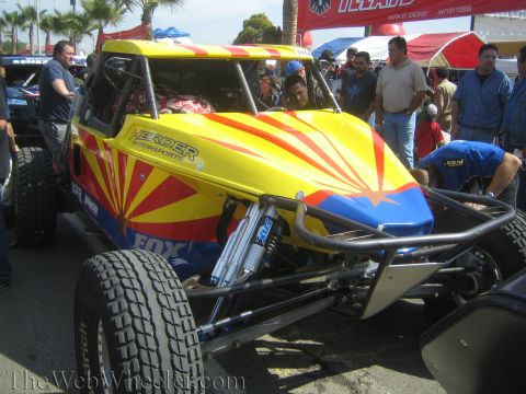 1990 Ford Bronco - brightly colored yellow, red and blue off-road race buggy with a sunburst graphic and exposed shock is parked among a crowd outdoors.