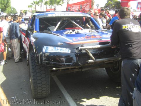 1990 Ford Bronco - black off-road race truck with Lucas sponsor graphics and a damaged front end is surrounded by onlookers at an outdoor event.