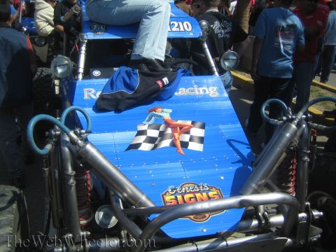 1990 Ford Bronco - blue rear deck of an off-road race car shows a pin-up girl with a checkered flag and a Genesis Signs decal, with someone seated above.