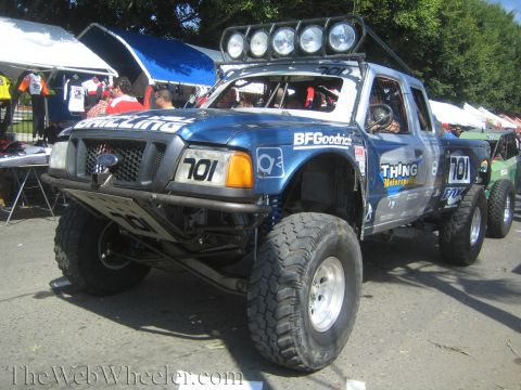 1990 Ford Bronco - blue Ford Ranger prerunner race truck numbered 701 with a roof-mounted light bar and BFGoodrich tires sits at an outdoor vendor area.