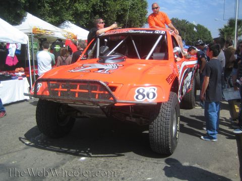 1990 Ford Bronco - orange off-road race truck numbered 86 with tribal graphics has two men seated atop it as a crowd gathers outdoors.