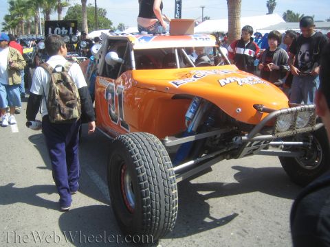 1990 Ford Bronco - orange single-seat off-road race buggy numbered 01 is parked on a street as spectators walk past at an outdoor event.