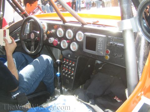 1990 Ford Bronco - Interior of a race car cockpit showing a carbon-fiber dash with round gauges, a steering wheel, a digital screen and a switch panel.