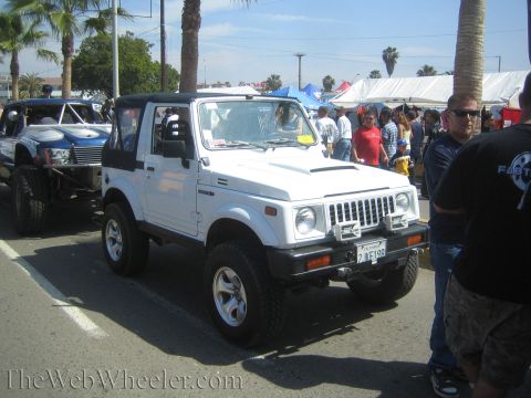 1990 Ford Bronco - white Suzuki Samurai 4x4 with a hardtop, lift kit and alloy wheels is parked on a street at a sunny outdoor event.