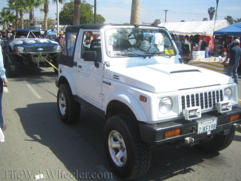 1990 Ford Bronco - white lifted Suzuki Samurai 4x4 with a California license plate is parked on a street near vendor stalls at an outdoor event.