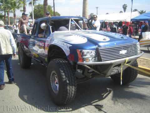 1990 Ford Bronco - blue Chevrolet off-road prerunner race truck numbered 66 with sponsor graphics is parked on a sunny street amid a crowd.