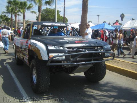1990 Ford Bronco - black-and-silver Chevrolet off-road race truck with a tube front bumper and big tires sits on a street at a busy outdoor event.