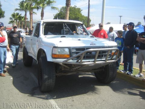 1990 Ford Bronco - white Ford prerunner race truck with a tube front bumper and off-road tires is parked on a street surrounded by spectators.
