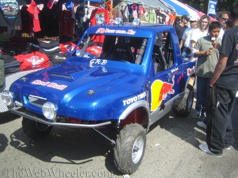 1990 Ford Bronco - blue off-road race truck wrapped in Red Bull and Toyo Tires graphics sits on display near a merchandise tent as spectators stand alongside.