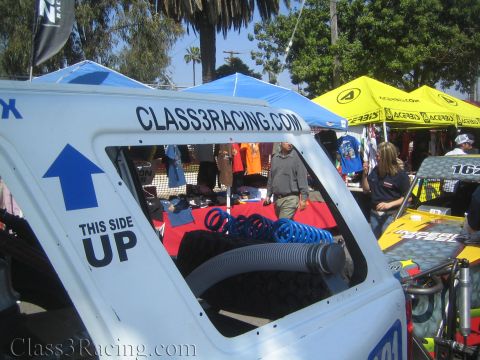 1990 Ford Bronco - white off-road race vehicle cab marked CLASS3RACING.COM and a This Side Up arrow stands in front of vendor tents and palm trees.