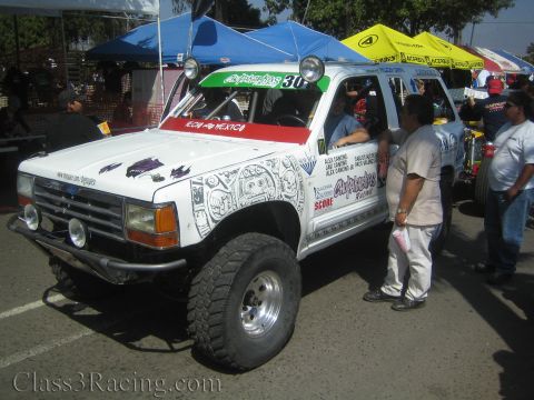 1990 Ford Bronco - white lifted Ford off-road race truck covered in detailed graphics sits under shade tents as people gather around it.