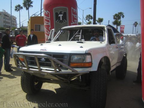 1990 Ford Bronco - white lifted Ford off-road truck with a tubular front bumper sits at a street event in front of a giant beer-can display.