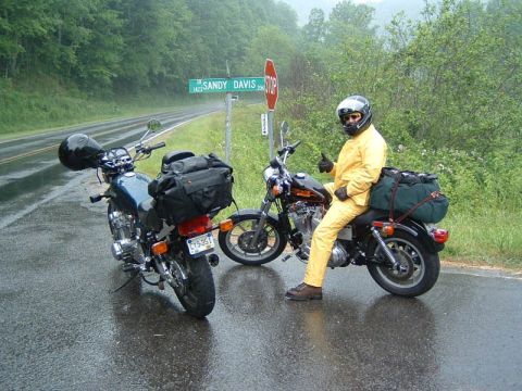 1985 Ford LTD Crown Victoria - A rider in yellow gear gives a thumbs up next to two motorcycles on a rainy day, with a street sign visible.