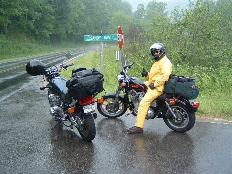 Two motorcycles parked on a wet road next to a Sandy Davis street sign. One rider in a yellow rain suit gives a thumbs up