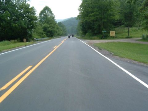 1985 Ford LTD Crown Victoria - Several motorcycles ride down a highway surrounded by lush green trees and rolling hills.