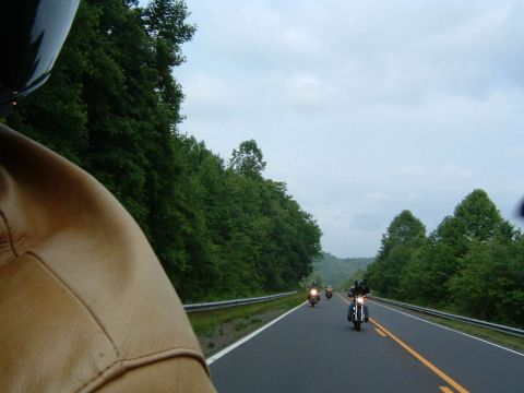 1985 Ford LTD Crown Victoria - A group of motorcycles travels down a two-lane highway surrounded by lush green trees under a cloudy sky.