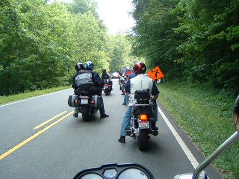 1985 Ford LTD Crown Victoria - A group of motorcyclists ride in formation on a scenic road, passing a temporary construction sign.