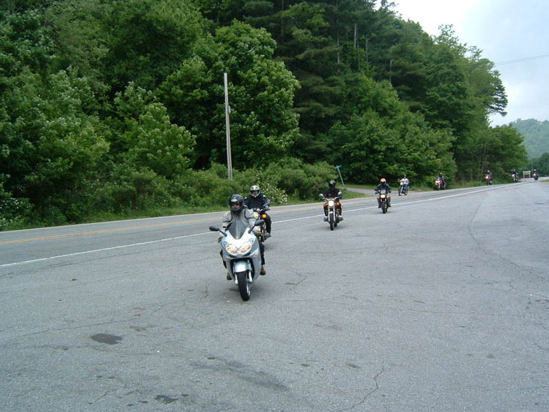 Motorcycles riding on a paved road with trees and a hill in the background