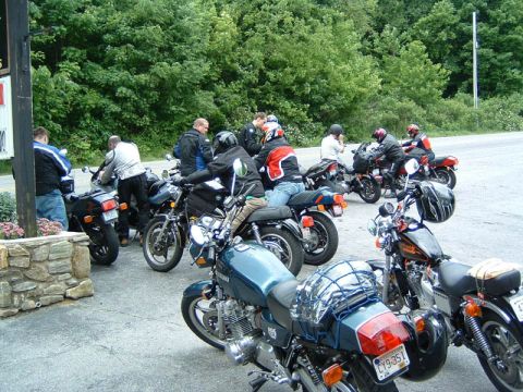 1985 Ford LTD Crown Victoria - A group of motorcyclists gather around their bikes on a paved area next to a wooded hillside.