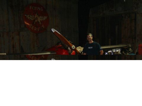 1985 Ford LTD Crown Victoria - A man stands beside a vintage red airplane with a large wooden propeller in a dimly lit hangar.
