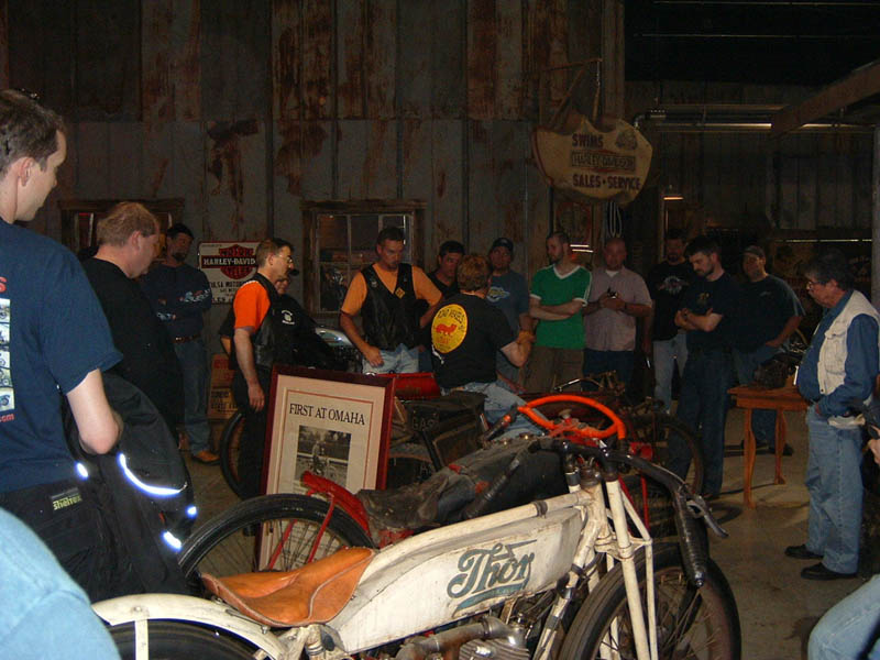 White Thor motorcycle with orange handlebars and brown seat, in a garage with people gathered around