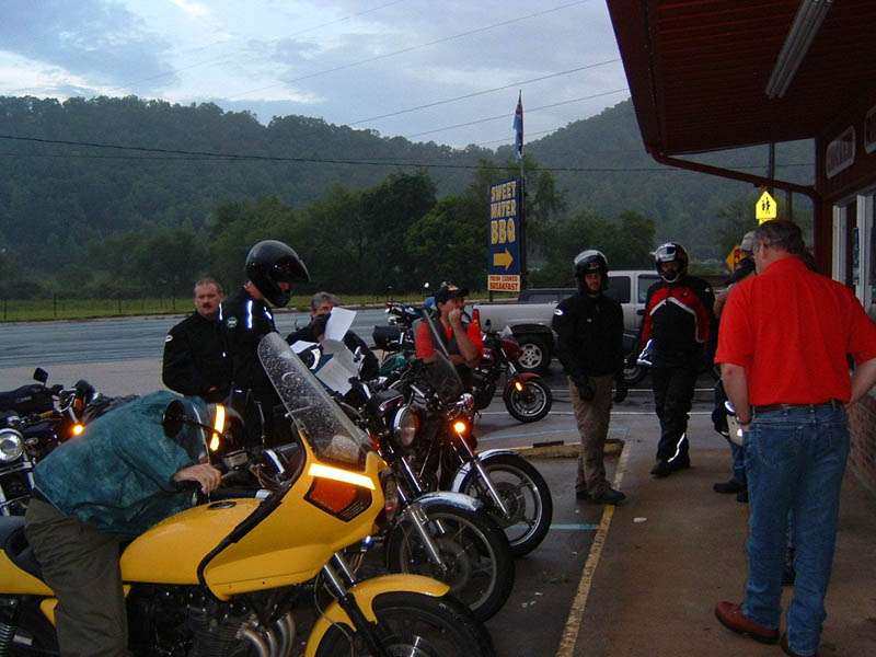 Yellow motorcycle in foreground, group of people and motorcycles in background, sign for Sweetwater BBQ