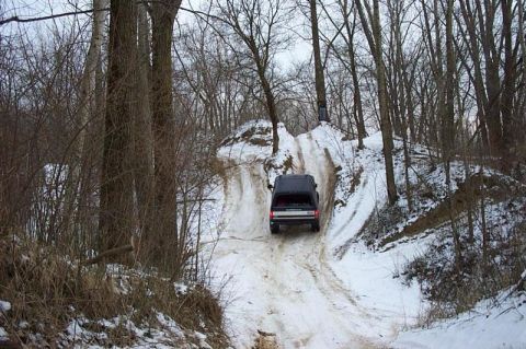 1992 Ford Bronco - Rear view of a dark full-size SUV climbing a steep, rutted hill of packed snow and ice on a wooded trail. Bare winter trees line both sides…