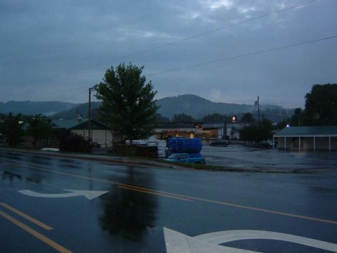 1985 Ford LTD Crown Victoria - A blue pickup truck carrying blue cargo is parked on a wet street in front of buildings under a cloudy sky.