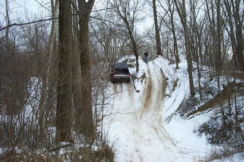 1992 Ford Bronco - Rear view of a dark 4x4 cresting a steep snow- and ice-covered hill climb cut through bare winter hardwoods, with rutted tire tracks runnin…