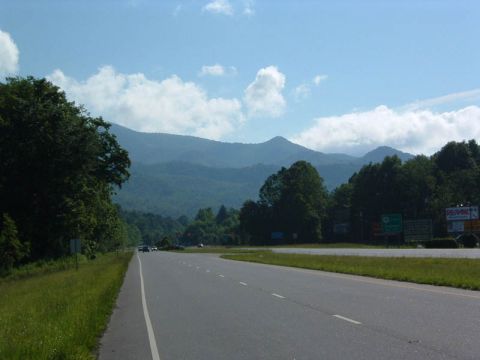 1985 Ford LTD Crown Victoria - A highway stretches towards distant mountains, with trees lining the roadside and clouds dotting the sky.