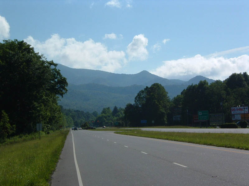 Highway passes by green trees and mountains under a cloudy blue sky. Signs are visible on the right