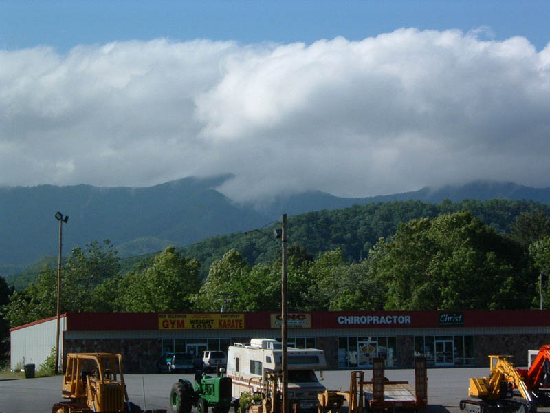 Yellow bulldozer, green tractor, and RV parked in front of a strip mall with mountains in the background
