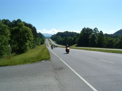 1985 Ford LTD Crown Victoria - A group of motorcycles cruises down a scenic highway on a clear day, surrounded by lush green trees and distant mountains.