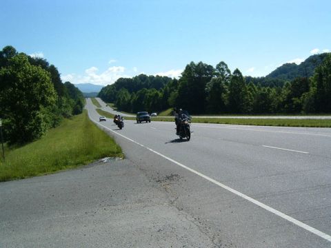 1985 Ford LTD Crown Victoria - Several motorcycles are cruising down a highway on a clear day, surrounded by lush green trees and distant mountains.