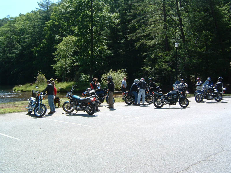 Motorcycles parked in a lot with people gathered, near water and trees